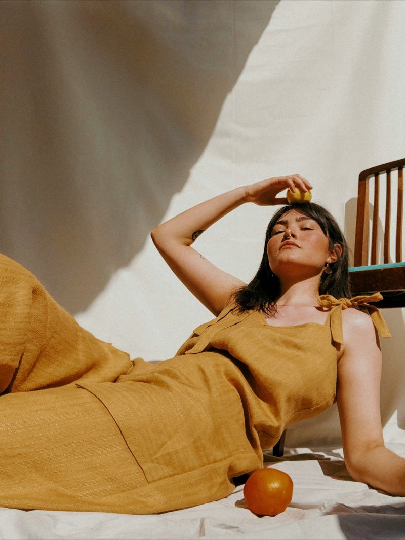 a woman in an ochre linen dress lying back in the sun, a lemon balanced on her head